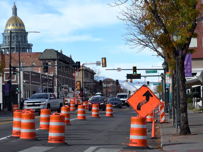 Road under construction with orange traffic cones and a right turn arrow sign, cars in the background, and a dome-shaped building visible in the distance.