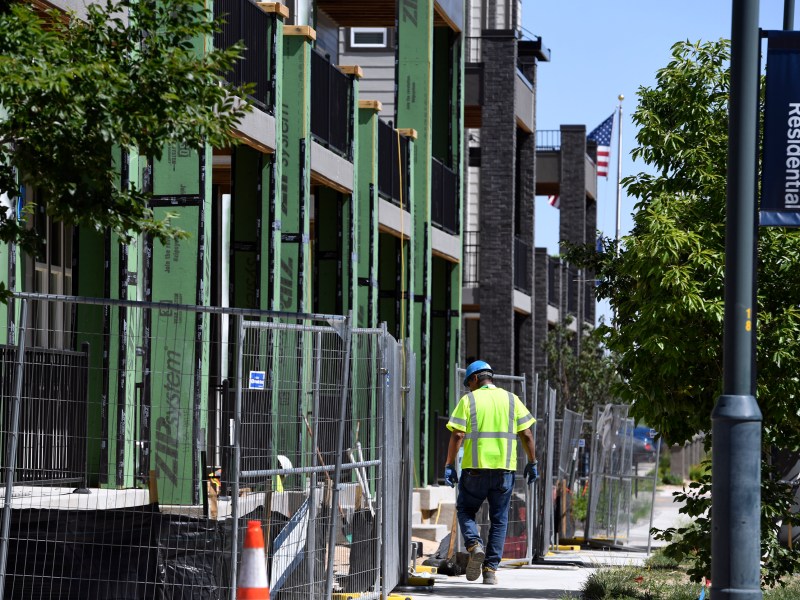 A man wearing a yellow safety shirt walks near fencing in front of townhomes that are under construction.