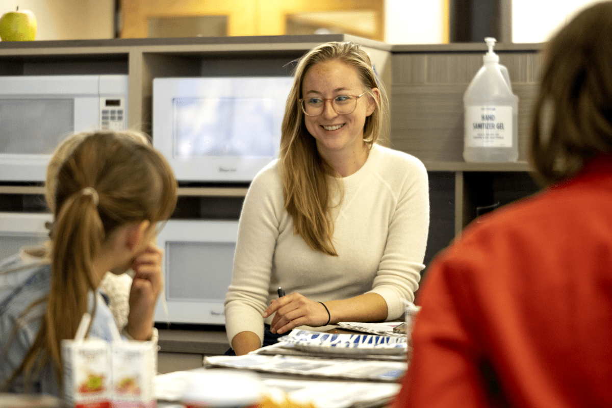 A woman with glasses sits at a table, smiling at students.